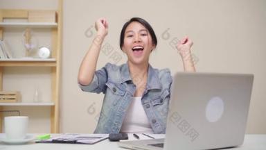年轻的亚洲妇女在家里客厅的办公桌上使用笔记本电脑工作。亚洲女商人成功庆祝在家庭办公室快乐跳舞的感觉。享受在家的时间概念.